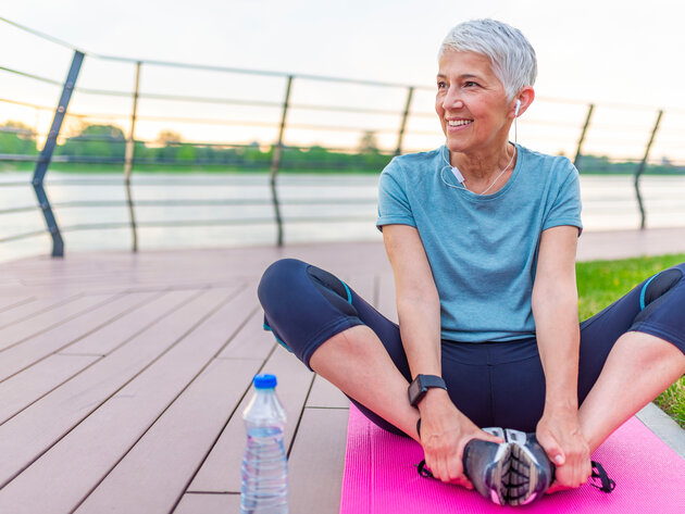 A smiling woman stands on a path in a park, holding a water bottle in one hand and a red yoga mat in the other. She is wearing a black athletic outfit and white headphones around her neck, surrounded by green trees and grass.