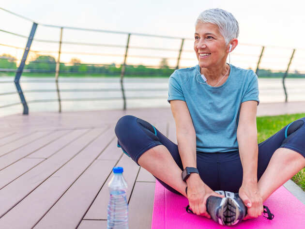 A smiling woman stands on a path in a park, holding a water bottle in one hand and a red yoga mat in the other. She is wearing a black athletic outfit and white headphones around her neck, surrounded by green trees and grass.