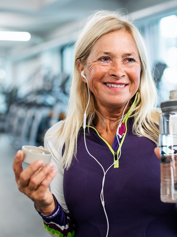 A smiling older woman with long blonde hair, wearing a purple athletic top and headphones, holds a water bottle in one hand and a small cup in the other. She stands in a gym with exercise equipment in the background.