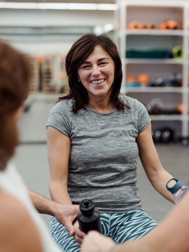 A smiling woman in a gray athletic shirt sits among friends in a fitness environment. She has medium-length dark hair and is engaged in conversation, holding a water bottle, with workout equipment visible in the background. The mood is friendly and casual.