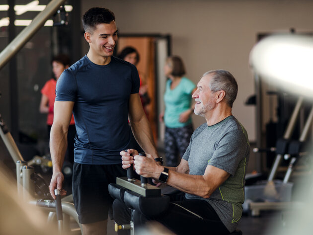 Two men are engaged in a fitness training session at a gym. One, a younger man in a blue shirt, is standing and smiling while assisting an older man seated at a workout machine. The older man has a content expression, suggesting a positive and supportive atmosphere.